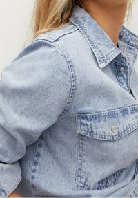 Veste en denim bleu clair avec un col pointu, des coutures visibles, une poche poitrine et des boutons-pression ; tissu texturé avec un aspect usé.