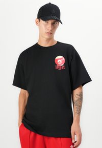 Sort bomulds-T-shirt med korte ærmer, prydet med et farverigt Adidas-logo på brystet. Bæres med røde bukser og en sort kasket.
