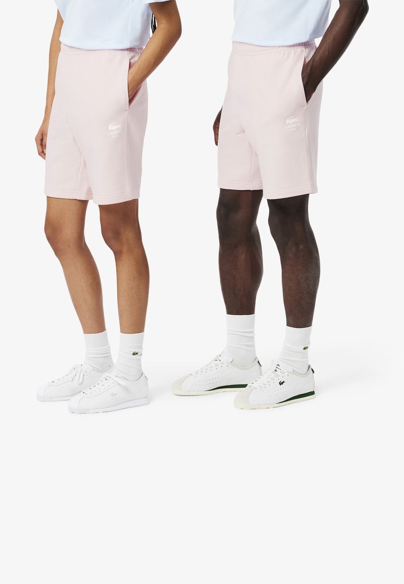 Shorts di cotone rosa chiaro con vita elasticizzata, tasche laterali e logo Lacoste stampato sulla gamba. Indossati con sneakers bianche e calze.