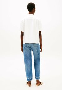 Camicetta bianca a maniche corte, vestibilità rilassata, con yoke sul retro. Abbinata a jeans azzurri a vestibilità ampia e scarpe slip-on beige. Sfondo semplice.