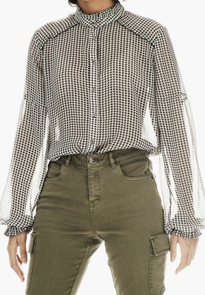 Person trägt eine schwarz-weiße Hahnentritt-Knopfbluse mit gepufften langen Ärmeln und olivgrünen Cargohosen.