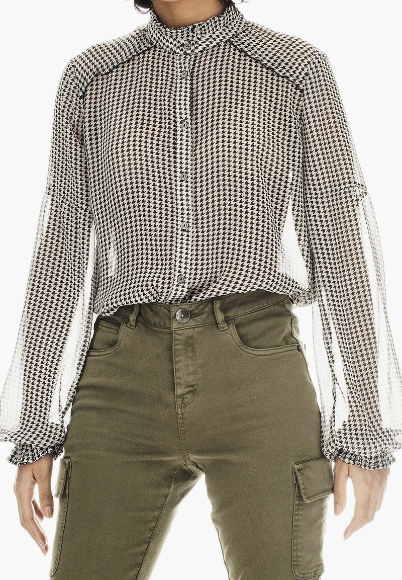 Personne portant un chemisier à boutons pied-de-poule noir et blanc avec manches longues bouffantes et un pantalon cargo vert olive.
