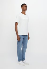 White short-sleeve T-shirt with textured logo on the front, paired with light blue straight-leg jeans and white sneakers on a neutral background.