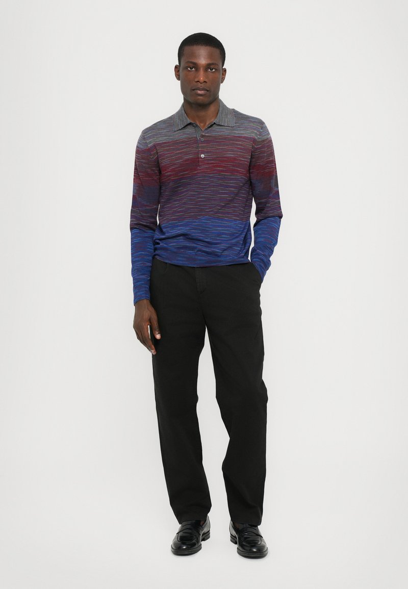 Camisa polo de manga larga con un patrón en degradado en tonos rojos, azules y grises; combinada con pantalones negros y zapatos negros.