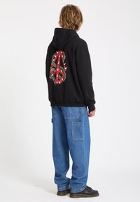 Sort hættejakke med et rødt og gyldent grafisk design på ryggen, parret med løst siddende blå denimbukser med lommer. Sorte sko.