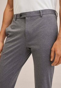 Close-up van een persoon in grijze op maat gemaakte broek met een knoop en riemlussen, hand rustend naast de dij.