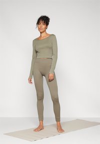 Maslinasto zelena rebrasta majica dugih rukava i odgovarajući uski tajice, s krojem koji je skraćen. Model stoji na svijetlom yoga prostirku.