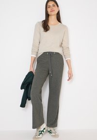 Femme portant un pull beige, un pantalon à cordon gris et des baskets blanches avec des détails verts, tenant une veste vert foncé à côté d'elle.