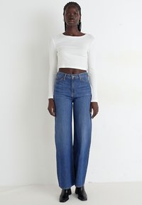 Top corto bianco a maniche lunghe abbinato a jeans blu a gamba larga con vita alta. Dettagli delle cuciture e chiusura classica con bottoni. Stivaletti neri alla caviglia.