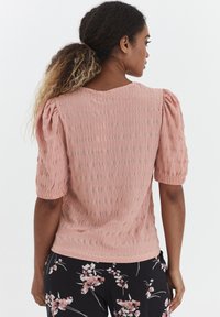 Top rosa chiaro con maniche a sbuffo realizzato in tessuto texturizzato, con scollo rotondo e vestibilità rilassata. Sfondo non visibile.