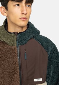 Fleecejack met bruine, groene en marineblauwe kleurblokken, een hoge kraag, rits sluiting en een logo tag aan de zoom. Zachte textuur.