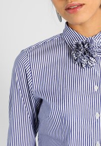 Blue and white striped shirt with a buttoned collar featuring a fabric flower detail at the neckline. Made from a lightweight, textured material.