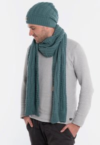 Zestaw niebiesko-zielonych dzianinowych czapki i szalika z teksturowanym wzorem, noszony na jasnoszarej bluzie. Szczegóły obejmują ściągacze i metkę.
