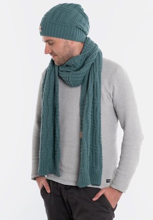 Teal gebreide muts en sjaal set met een gestructureerd patroon, gedragen over een lichtgrijze trui. Details omvatten ribbelranden en een label.