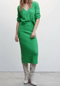 Grünes gestricktes zweiteiliges Outfit mit einem V-Ausschnitt-Pullover und einem figurnahen Midi-Rock, kombiniert mit hohen, weißen, Absatzstiefeln. Texturiertes Muster auf dem gesamten Kleidungsstück.