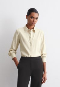 Cămașă galben deschis cu guler, cu mâneci lungi răsucite și nasturi în față, asortată cu pantaloni negri cu talie înaltă. Textură de țesătură netedă.