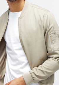 Chaqueta bomber beige con puños y cuello acanalados, que presenta un bolsillo con cremallera en la manga y una textura suave y ligera.