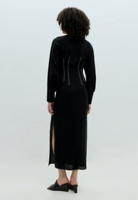Schwarzes langärmeliges Kleid mit einem seitlichen Schlitz, das kontrastierende weiße Nähte auf dem Rücken aufweist. Aus strukturiertem Stoff gefertigt. Getragen mit schwarzen Absatzsandalen.