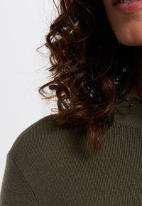 Olijfgroene gebreide trui met ribbelstructuur, met een hoge hals en versieringen op de schouder. Losse, krullende bruine haren zijn zichtbaar.
