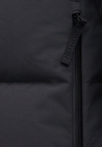 Geacă puffer neagră dintr-un material mat, cu închidere prin fermoar și detalii de umplutură cosute. Eticheta de tragere a fermoarului este vizibilă pe lateral.