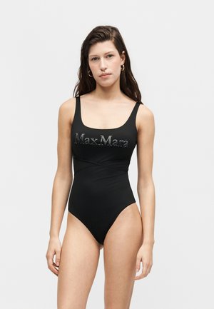Fiatal nő hosszú sötét hajjal, fekete ujjatlan bodyt visel, rajta a "Max Mara" logóval, egyszerű világos háttér előtt áll.