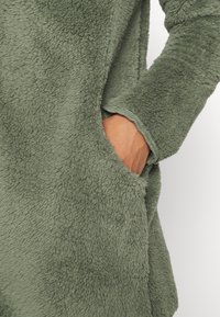 Green fleece garment with a soft, textured surface, featuring a side pocket and loose-fitting sleeves.