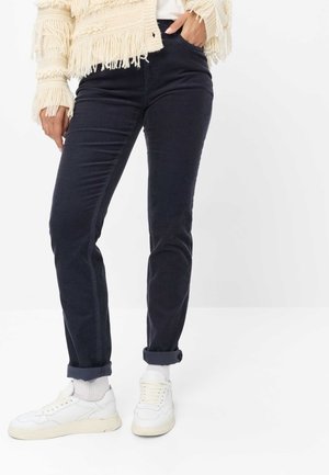 Personne portant un pantalon foncé retroussé, des baskets blanches, des chaussettes blanches et un pull beige à franges, une main dans la poche.