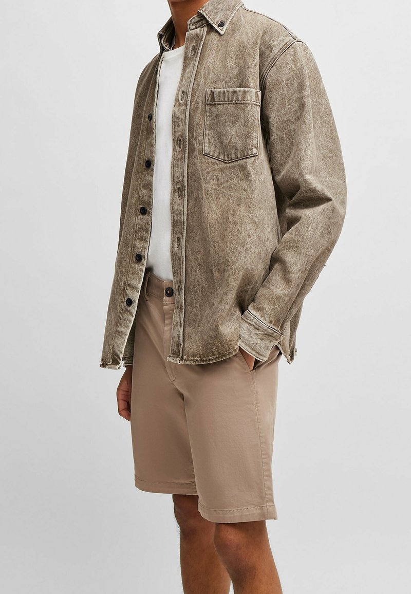 Homme portant une chemise boutonnée marron délavée sur un t-shirt blanc avec un short beige arrivant aux genoux, debout avec une main dans la poche.