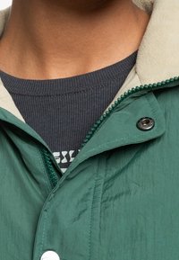 Lekka zielona kurtka z kremowym kołnierzem, zapinana na zatrzaski, z kontrastowym zamkiem błyskawicznym zdobionym koralikami. Pod spodem widoczna jest szara teksturowana koszulka z okrągłym dekoltem.