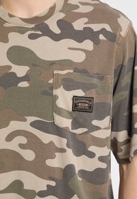 Camo-gepatroneerde T-shirt in aardetinten met een zak op de borst en een label. Gemaakt van zacht katoen, met korte mouwen.