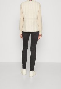 Beżowa pikowana kamizelka z długimi rękawami, kontrastowym kremowym materiałem na rękawach i ściągaczami przy mankietach. Noszona na ciemnych obcisłych dżinsach i białych trampkach.