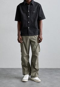 Camicia nera a maniche corte con bottoni e tasca sul petto, abbinata a pantaloni cargo verde oliva e sneaker bianchi. Design semplice con tessuto traspirante.