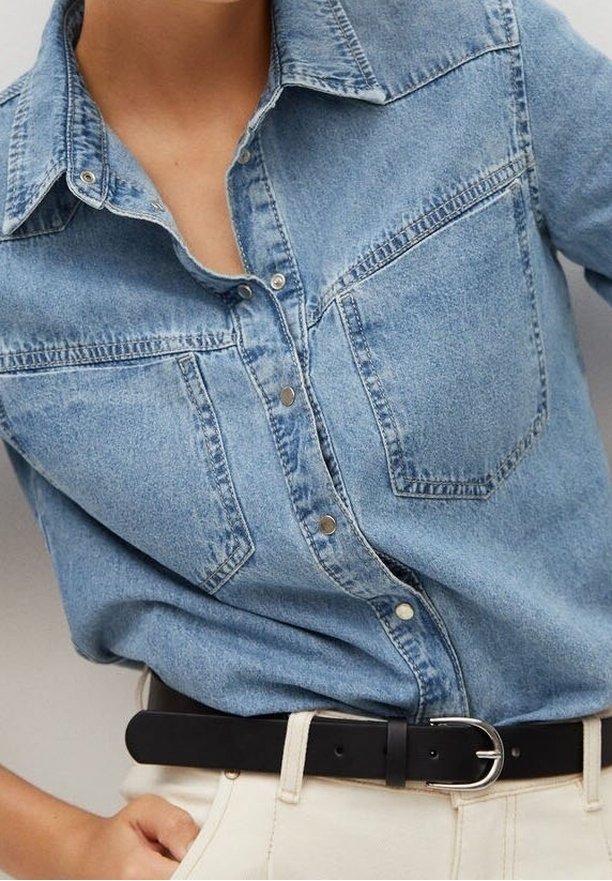 Femme portant une chemise en denim bleu clair avec des boutons-pression, rentrée dans un pantalon beige clair avec une ceinture noire à boucle argentée.