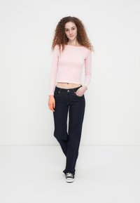 Ανοιχτό ροζ crop top με μακριά μανίκια, συνδυασμένο με σκουρό μπλε τζιν παντελόνι ίσιας γραμμής. Το μοντέλο φοράει μαύρα αθλητικά παπούτσια. Ομαλή υφή με ελάχιστες λεπτομέρειες.