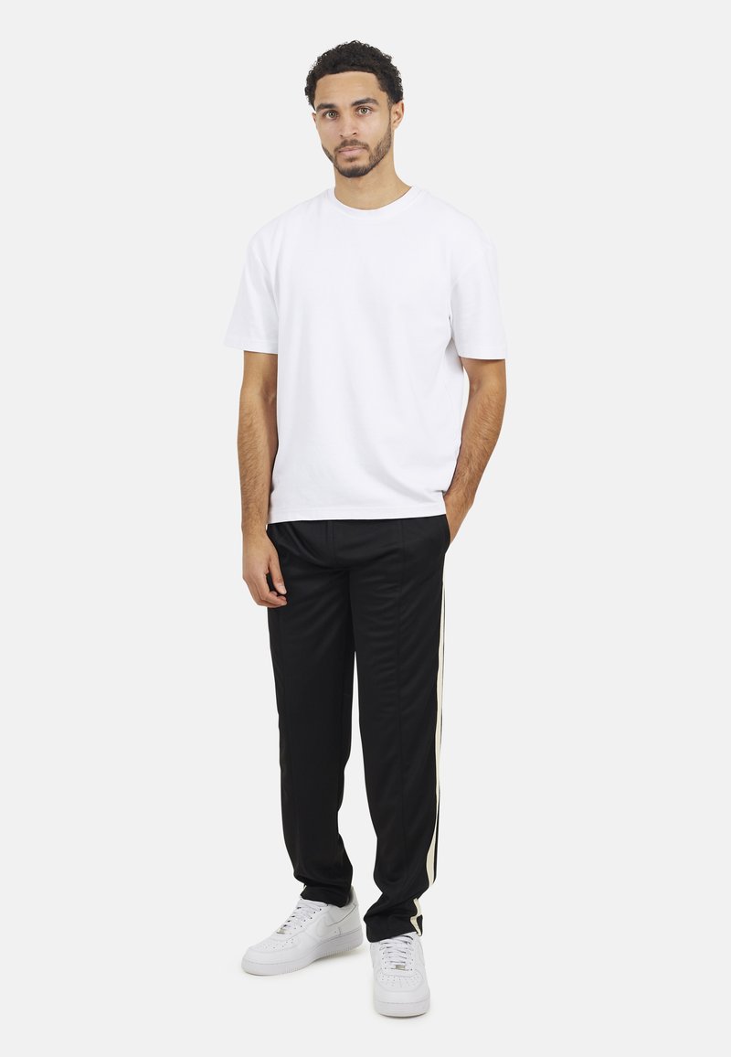 Homme debout avec les mains dans les poches, portant un t-shirt blanc, un pantalon de jogging noir avec une bande sur le côté et des baskets blanches, sur fond blanc.