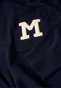 Tissu tricoté bleu marine foncé avec une grande lettre M blanche brodée et un poignet de manche côtelé visible.