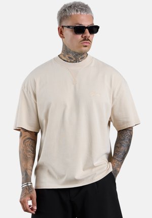 Mann mit hellgrauen Haaren und Tattoos, der schwarze Sonnenbrille, beige übergroßes T-Shirt und schwarze Hose vor einfarbigem Hintergrund trägt.