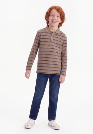 Garçon roux souriant debout, portant un polo à manches longues marron à rayures, un jean bleu et des baskets blanches sur fond blanc.