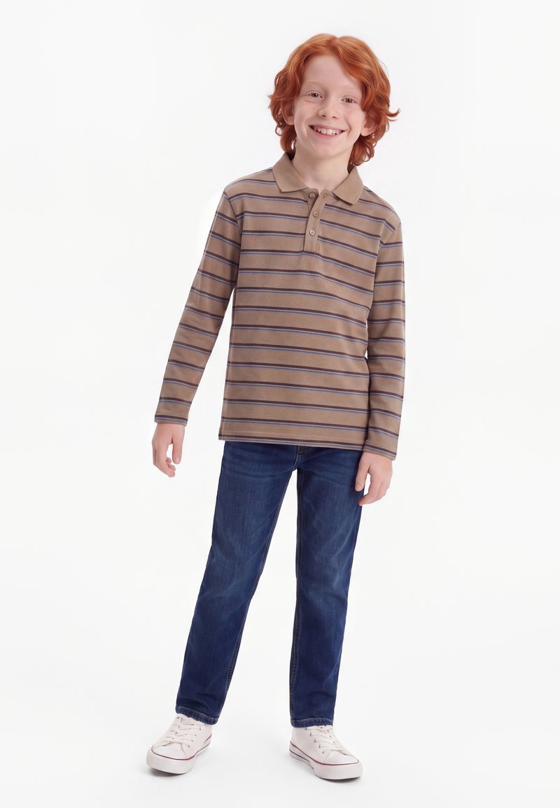 Garçon roux souriant debout, portant un polo à manches longues marron à rayures, un jean bleu et des baskets blanches sur fond blanc.
