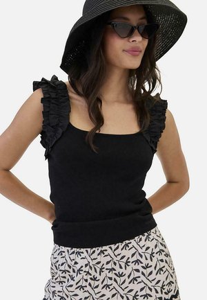 Mujer con blusa negra sin mangas con volantes, sombrero negro de ala ancha, gafas de sol estilo ojo de gato y falda blanca con estampado de hojas negras, posando con las manos en las caderas.