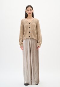 Beżowy dziergany kardigan z dekoltem w kształcie litery V, w zestawieniu z wysokim stanem, plisowanymi srebrnymi spodniami. Czarne guziki ozdabiają przód kardiganu.