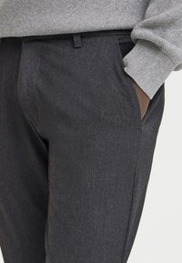 Temno sivi hlače iz teksturirane tkanine z ozkim krojem, ravnim sprednjim delom in stranskimi žepi. Obute z svetlo sivim puloverjem.