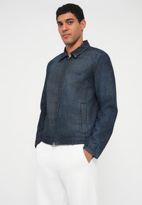 Τζιν μπουφάν σε σκούρο μπλε με υφασμάτινο μοτίβο, φερμουάρ μπροστά, μυτερός γιακάς και δύο πλαϊνές τσέπες. Άσπρο παντελόνι που φοριέται από κάτω.