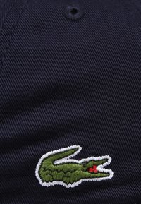 Cască din bumbac de culoare bleu marin, având un logo brodat cu un crocodil verde și un accent roșu. Formă structurată și detalii de coasere vizibile.