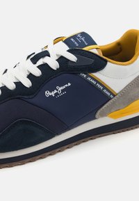 Meriinisininen lenkkari valkoisilla ja keltaisilla yksityiskohdilla, jossa on sekoitus mokkaa ja tekstiilimateriaaleja, litteät valkoiset nauhat ja teksturoitu kumipohja.