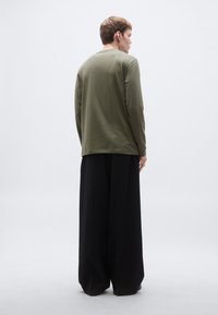 Μακρυμάνικο πουκάμισο σε ελιά πράσινο με άνετη εφαρμογή, συνδυασμένο με μαύρο παντελόνι φαρδιάς γραμμής, εμφάνιση από πίσω πάνω σε απλό φόντο.