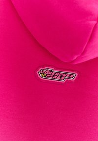 Jasně růžová tkanina obsahuje odznak s textem "THE POWERPUFF GIRLS" a grafické prvky, našité na materiálu. Hladký povrch.