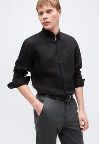 Νέος άνδρας με ανοιχτόχρωμα μαλλιά, φοράει μαύρο πουκάμισο με μακριά μανίκια, κουμπωτό με ρολά μανίκια και γκρι παντελόνι με μαύρη ζώνη, στέκεται με το ένα χέρι στην τσέπη.