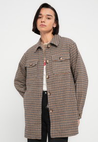 Καρό παλτό σε καφέ και κρεμ αποχρώσεις με κουμπιά μπροστά, δύο τσέπες στο στήθος και άνετη εφαρμογή, με γιακά και υφασμάτινη υφή.