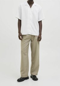 Uomo che indossa una camicia bianca a maniche corte con bottoni, pantaloni beige a gamba larga e scarpe di pelle nere su uno sfondo neutro.
